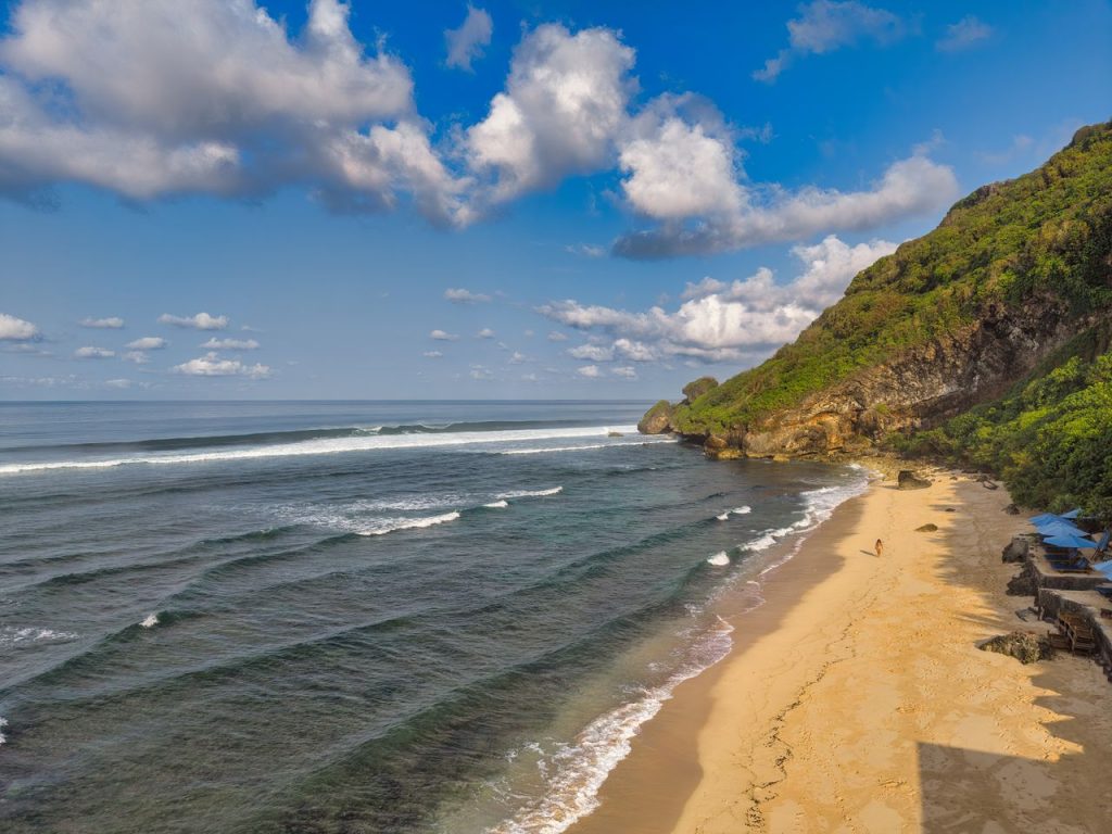 Explore the serene beauty of a Bali beach with azure waters, golden sands, and lush cliffs under a bright sky.
